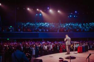 A speaker in a gray suit captivates a large crowd on stage during a motivational event.